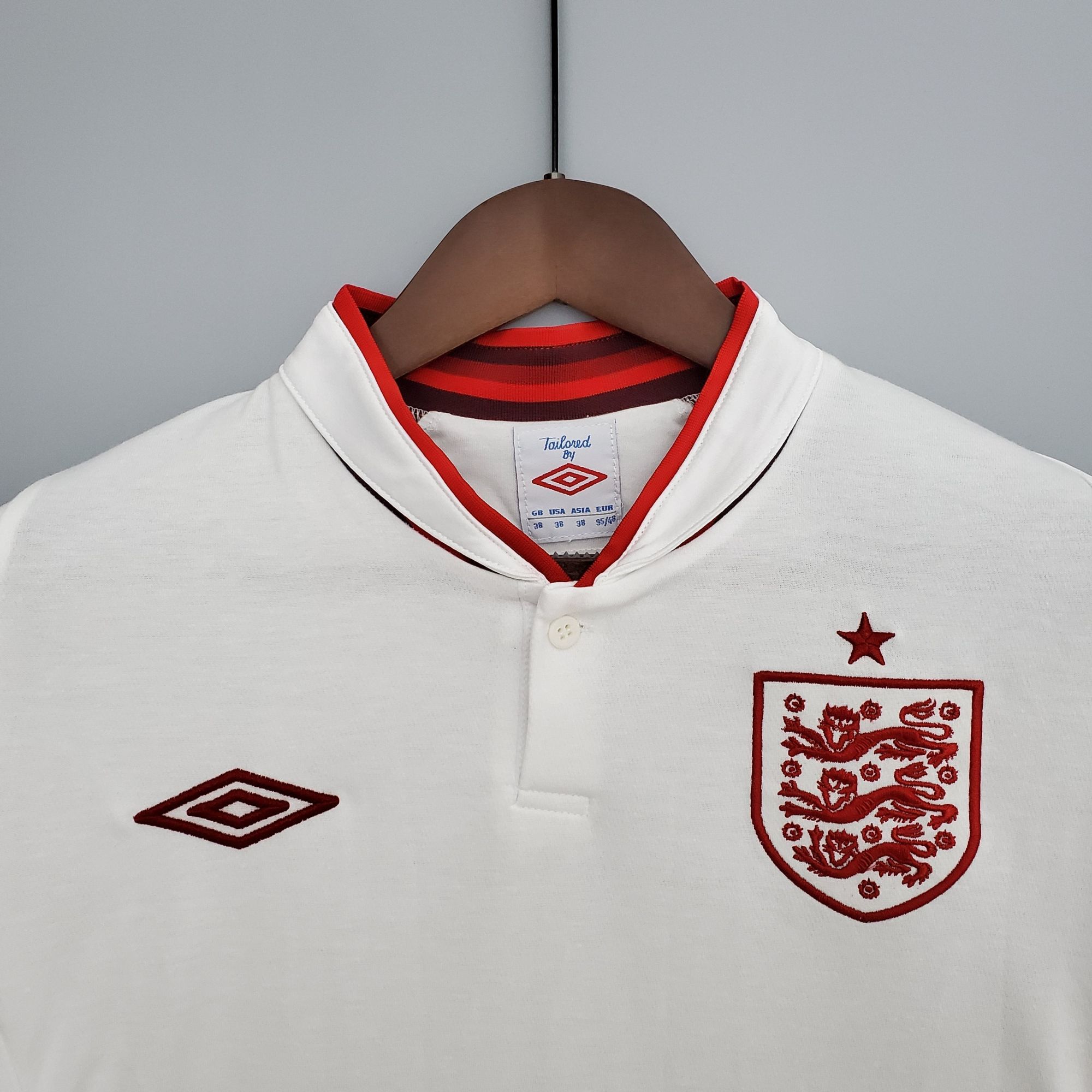 56181139_fe4dcc91-f0cf-4a1e-aa43-dfe7a876323a | UAE SPORT STORE White polo shirt on a wooden hanger with red trim at the neckline and a red crest on the chest, hanging against a gray background.