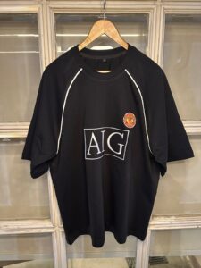 Black Manchester United-themed sports shirt on a wooden hanger, with white piping and the AIG logo on the chest and a small club crest nearby.