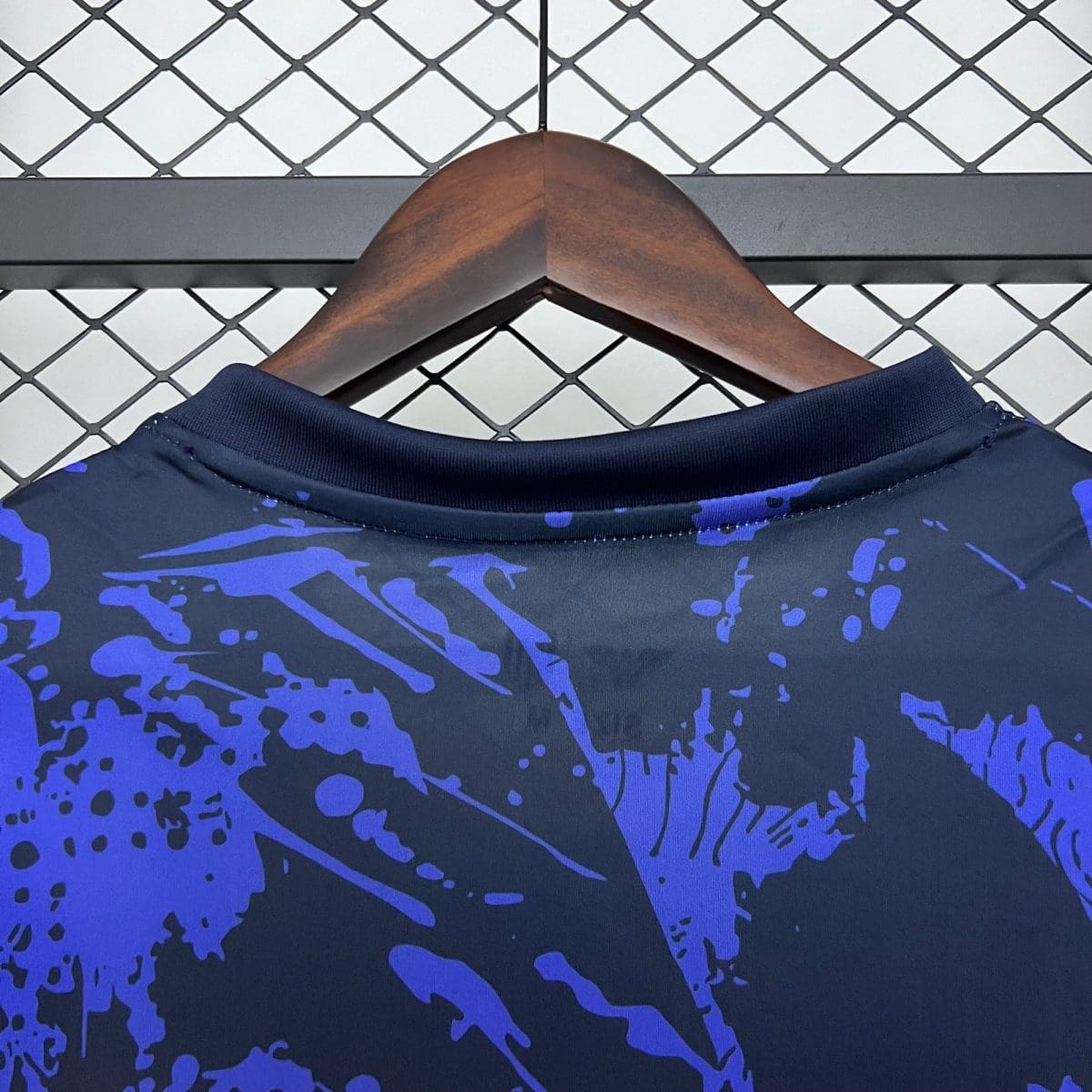 Close-up of a wooden clothes hanger holding a dark blue shirt with purple/blue splatter, against a chain-link fence background.