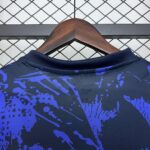 Close-up of a wooden clothes hanger holding a dark blue shirt with purple/blue splatter, against a chain-link fence background.