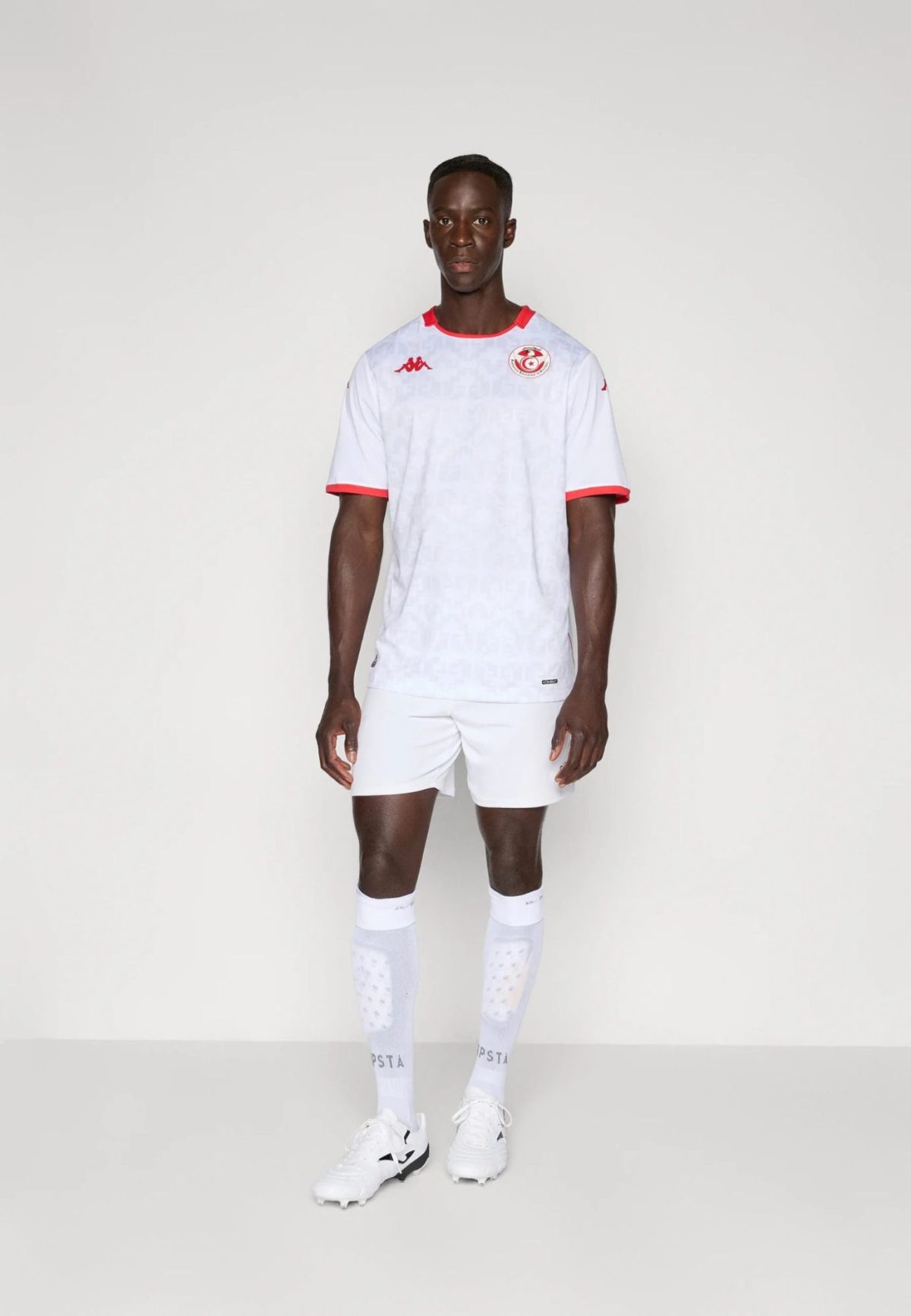 Male soccer player standing in a white kit with red trim, white shorts and socks, against a light background.