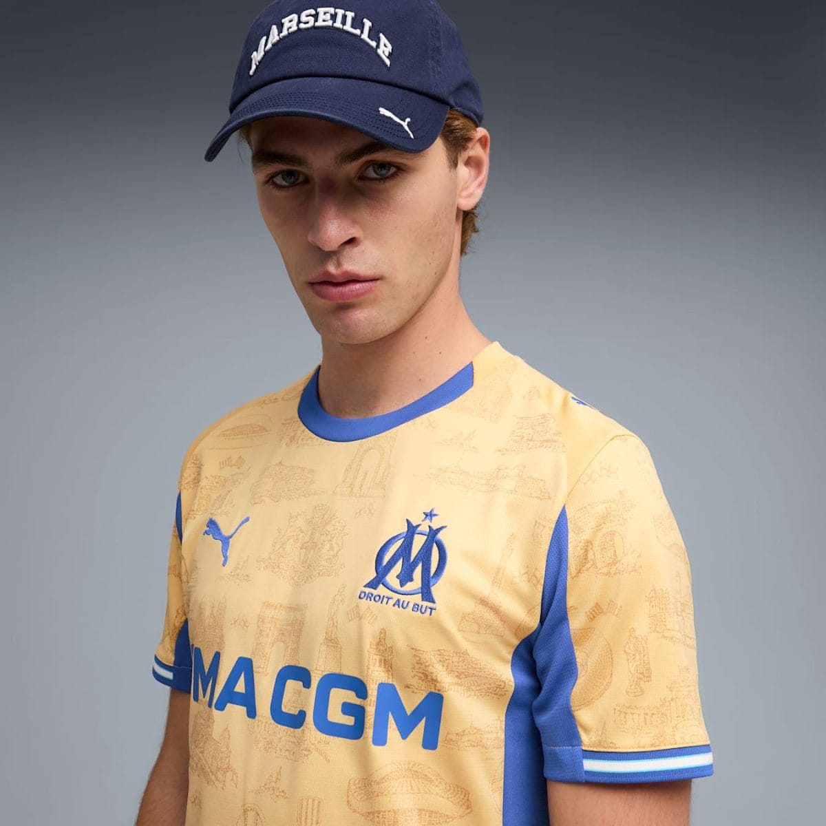 Portrait of a young man wearing a blue MARSEILLE cap and a pale yellow OM soccer jersey with blue trim and print details.