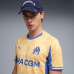 Portrait of a young man wearing a blue MARSEILLE cap and a pale yellow OM soccer jersey with blue trim and print details.