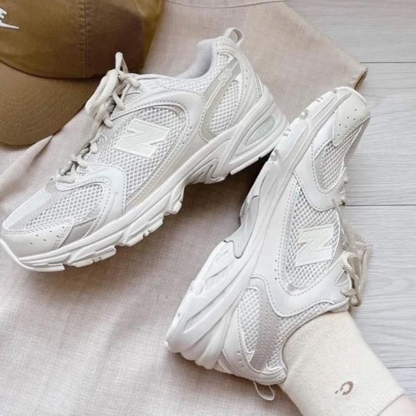 White chunky athletic sneakers with mesh panels, resting on light beige fabric with a brown cap nearby.