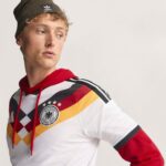Young man modeling a Germany national football jersey with red, black, and gold argyle patterns and a circular crest on the chest, looking to the left.