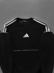 Black Adidas sweatshirt with white stripes along the sleeves and a white Adidas logo on the chest, laid flat on a gray surface.