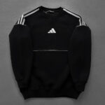 Black Adidas sweatshirt with white stripes on shoulders and logo, laid flat on a gray background.