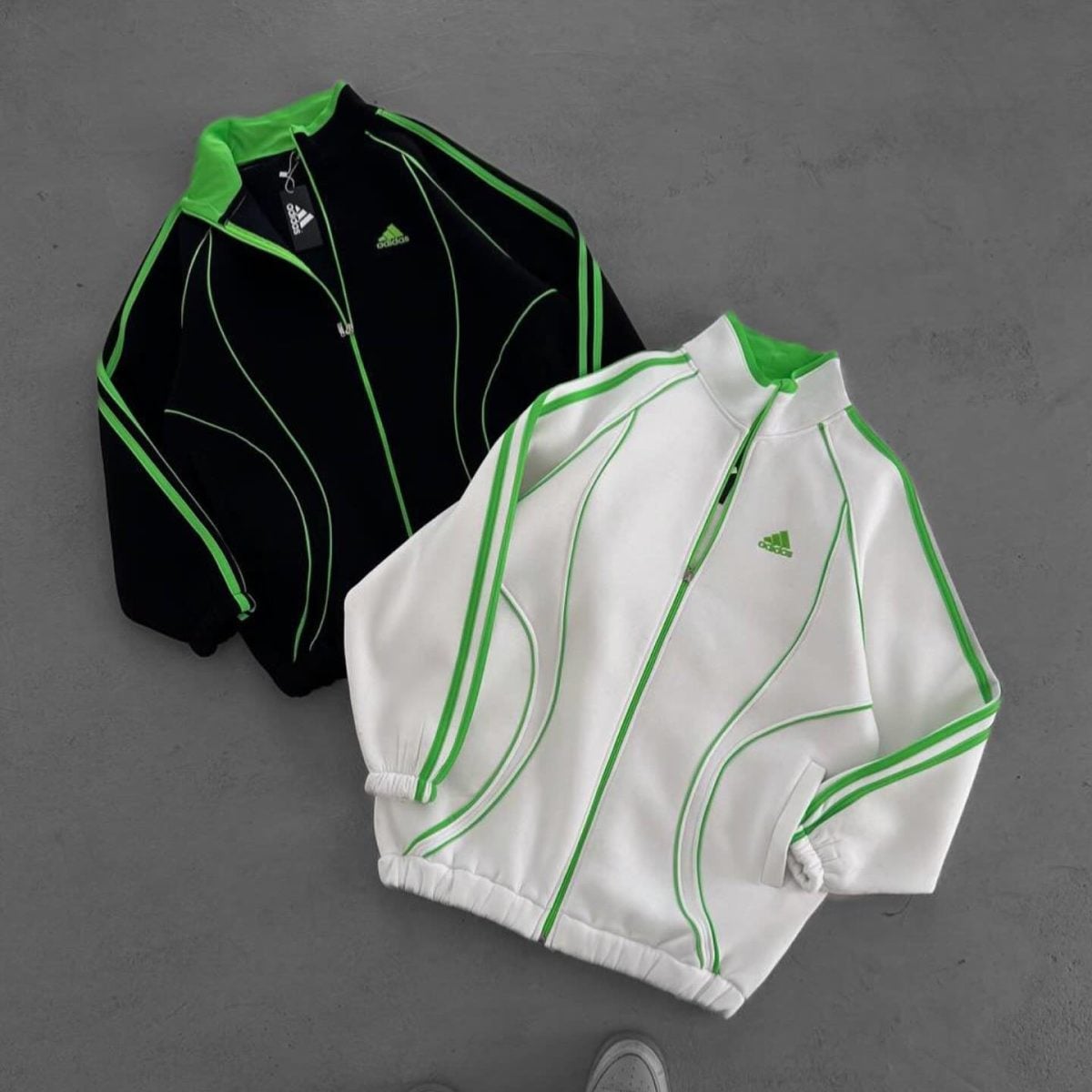Two Adidas track jackets (black and white) with neon green piping laid on a gray surface.