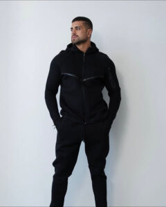 Man in a black zip-up hoodie and matching joggers stands with hands in pockets, looking to his left against a pale wall.