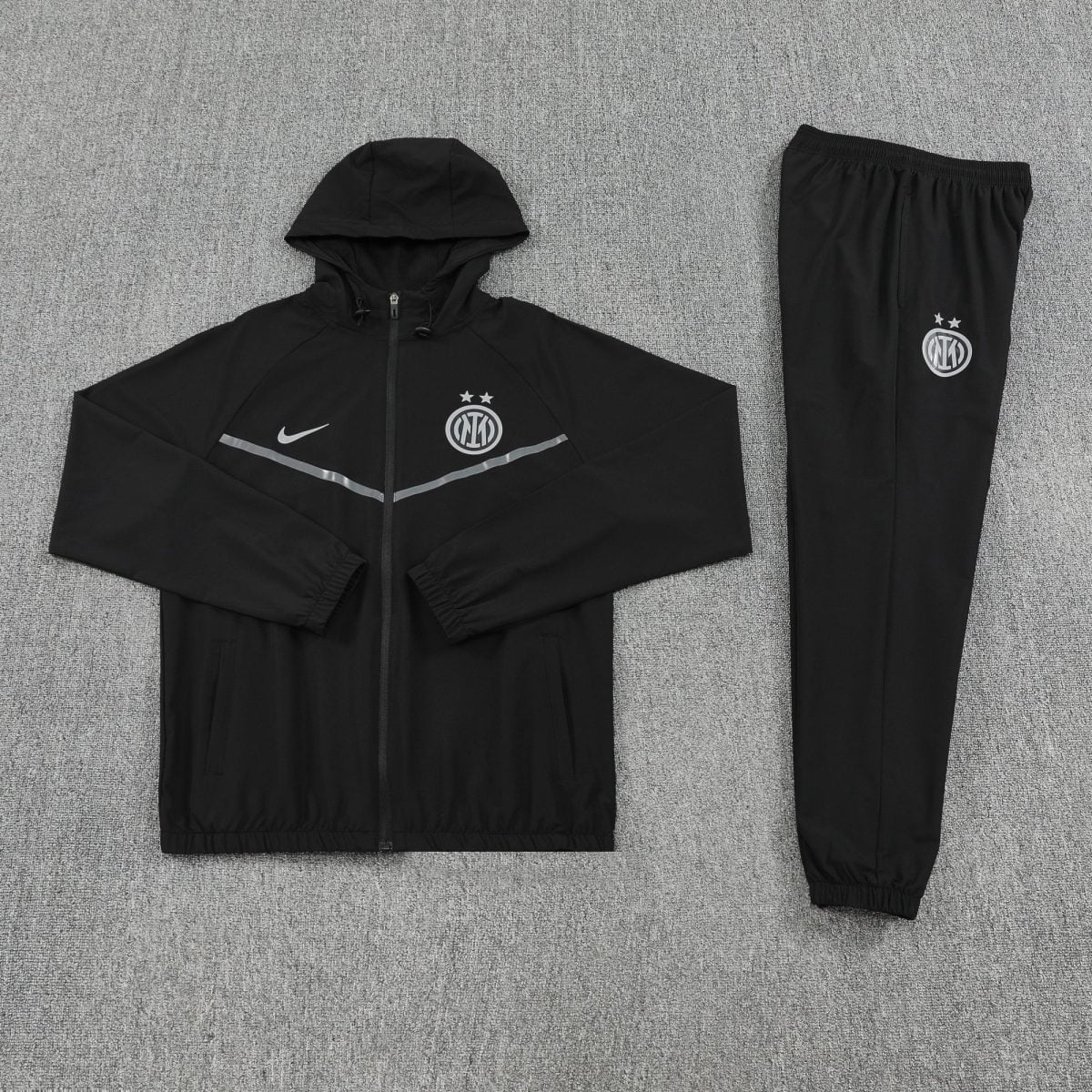 Black Inter Milan tracksuit laid flat: hooded zip-up jacket with crest and a matching pair of joggers on a gray carpet surface.