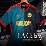 57hzbwJ20EMI6mP | UAE SPORT STORE LA Galaxy retro-style soccer jersey on a hanger with vintage equipment in the background, blue and black with red accents.