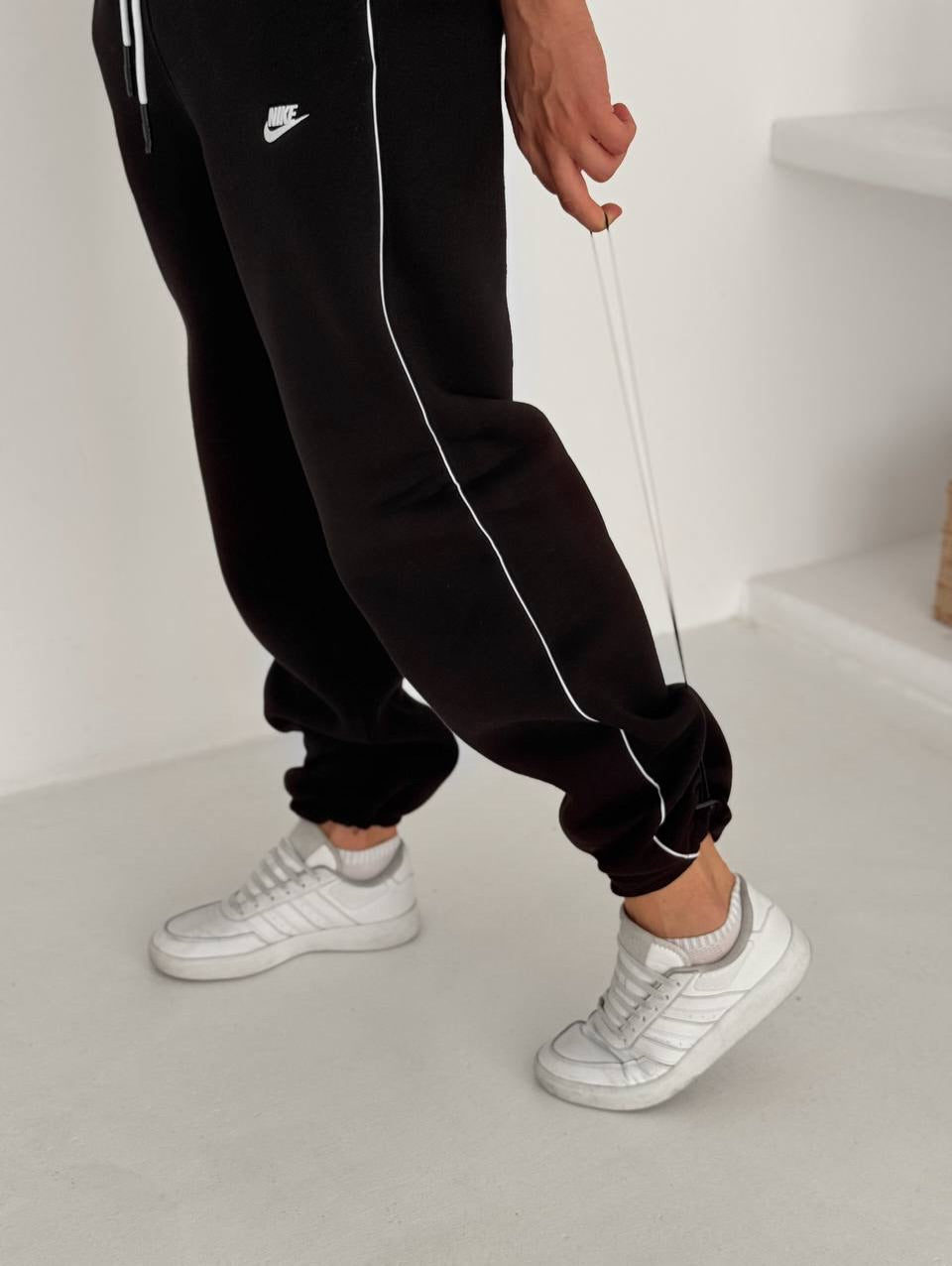 586669538_17891230320372975_1445268283140170138_n | UAE SPORT STORE Person wearing black joggers with white piping and a small Nike logo, paired with white sneakers, stepping indoors.