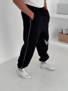 586687607_17891230293372975_7050443102693213103_n | UAE SPORT STORE Person wearing black joggers with a white side stripe and a silver Nike swoosh, paired with white sneakers, standing in a bright, minimalist room.