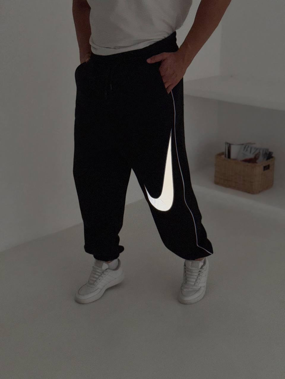 586933518_17891230302372975_2756201093738462569_n | UAE SPORT STORE Person standing indoors in black joggers with a white Nike swoosh stripe down the side, wearing white sneakers and a white shirt.