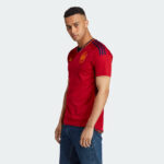 Man modeling a red Spain national football team jersey with navy stripes on the shoulders and Adidas logo, standing in profile.
