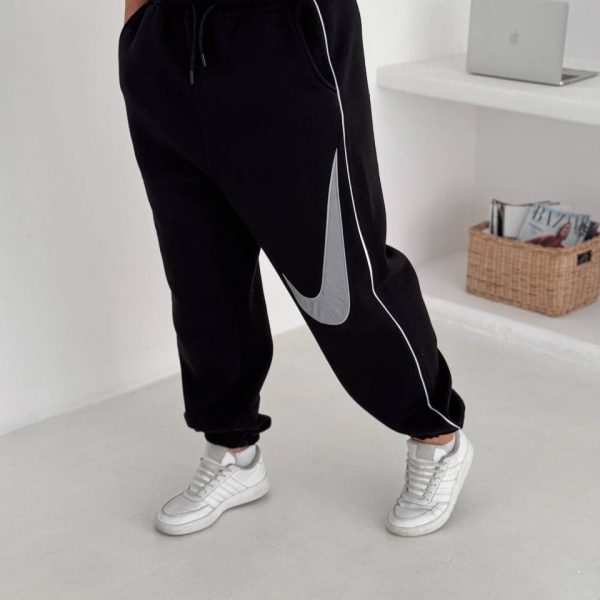 Person wearing black joggers with white piping and a large gray Nike swoosh, paired with white sneakers, standing in a bright room.