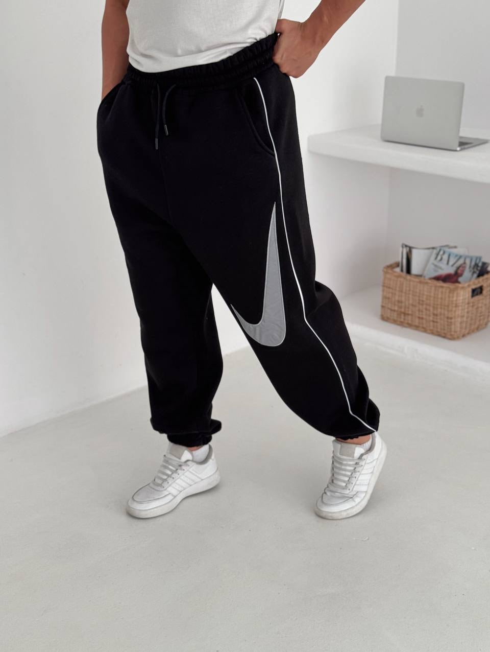 587274019_17891230311372975_4237086960217873955_n | UAE SPORT STORE Person wearing black joggers with white piping and a large gray Nike swoosh, paired with white sneakers, standing in a bright room.