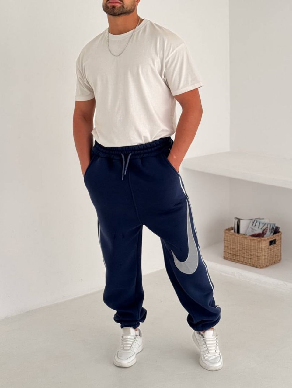 587277030_17891230347372975_3757599815386115145_n | UAE SPORT STORE Man in a white t-shirt and navy joggers with a large gray Nike swoosh, white sneakers, standing with hands in pockets in a bright room.