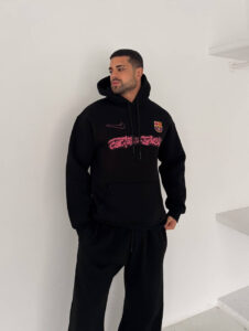 Man in a black hoodie with a Barcelona crest, posing against a white wall with hands in pockets.