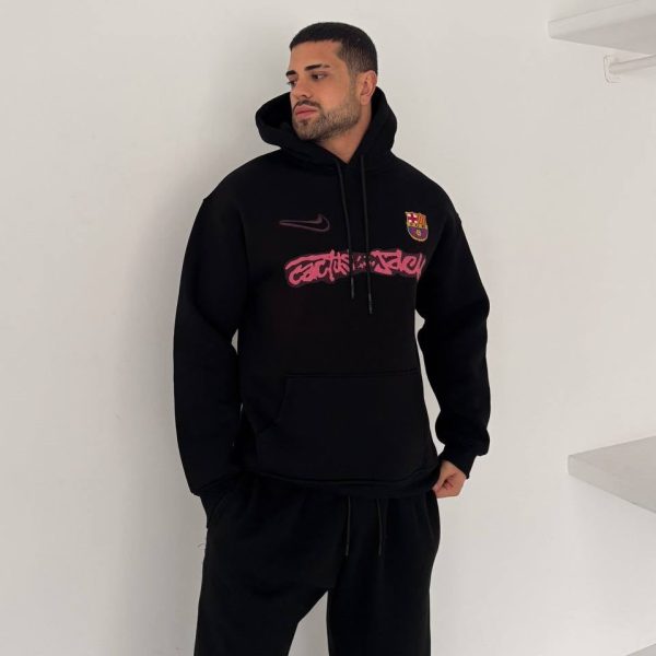 Man in a black hoodie with a Barcelona crest, posing against a white wall with hands in pockets.