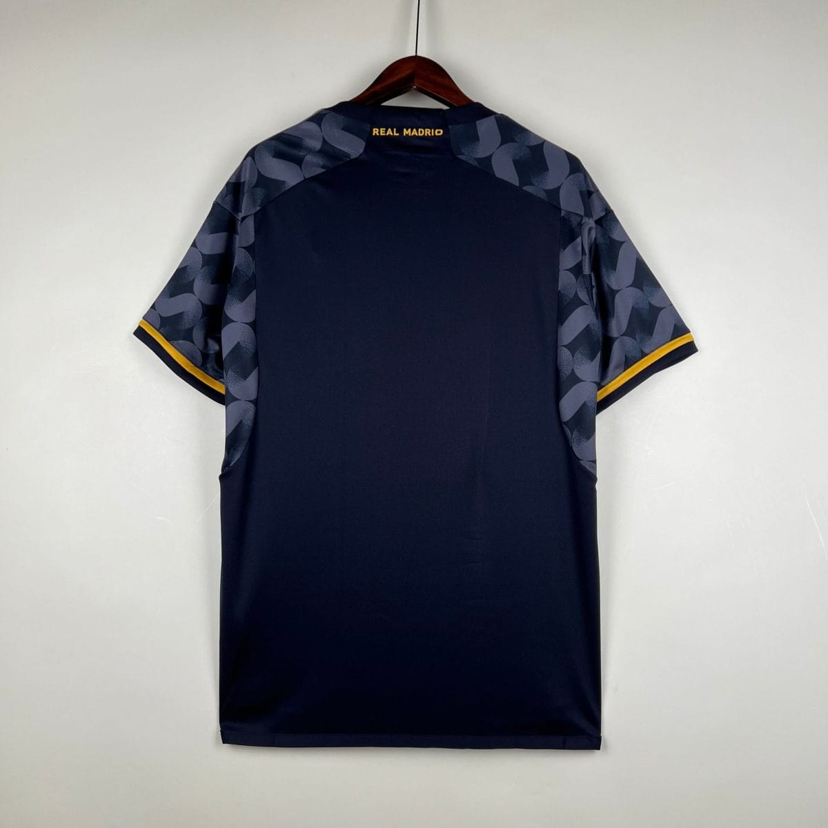 Navy shirt on a wooden hanger, back view with blue patterned sleeves, yellow trim at the cuffs, and 'REAL MADRIO' in gold on the neck area.