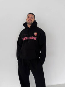 Man wearing a black hoodie with a Barcelona crest and pink graffiti text, standing against a light gray wall.