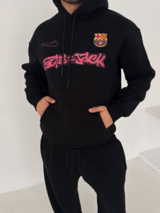 Person wearing a black hoodie with FC Barcelona crest and pink graffiti-style text, hands in pockets, standing indoors.