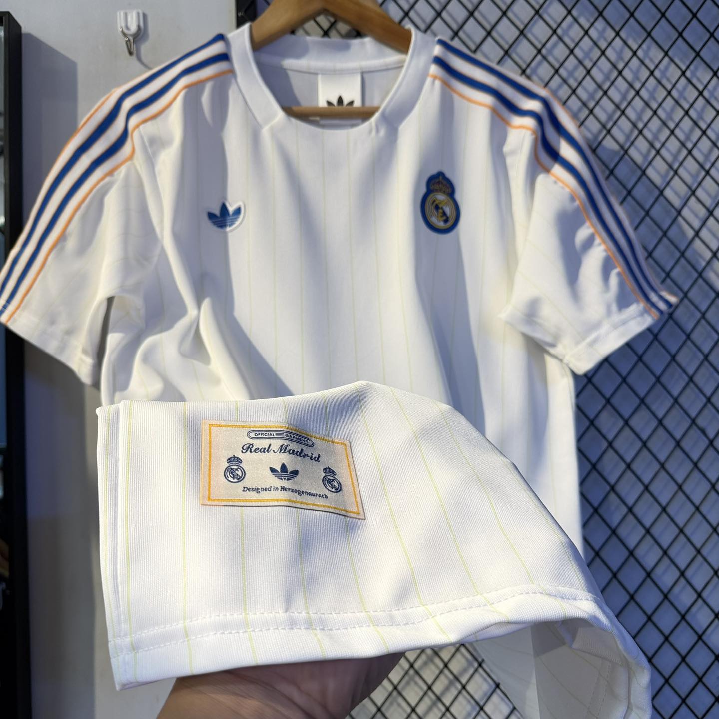 593723164_1158609296421624_4339424859135861567_n | UAE SPORT STORE White Real Madrid soccer jersey with blue/orange shoulder stripes, hanging on a wooden hanger.