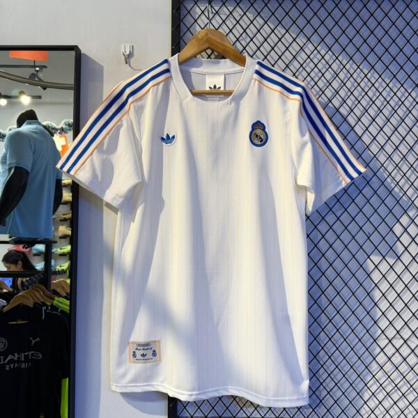 White Real Madrid soccer jersey with blue Adidas stripes on the shoulders and Real Madrid crest, hanging on a wooden hanger.