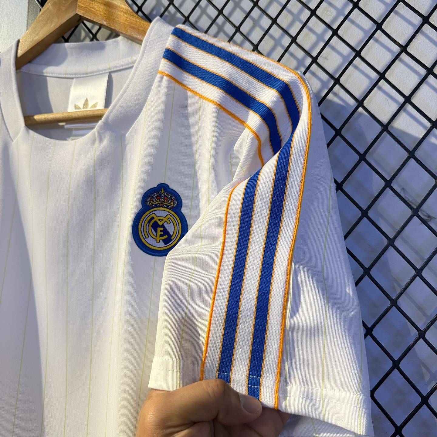 595162909_1158609289754958_6733636137515774258_n | UAE SPORT STORE White Real Madrid jersey with blue and orange striped shoulder detail and crest on chest, hanging on a wooden hanger in front of a geometric fence.