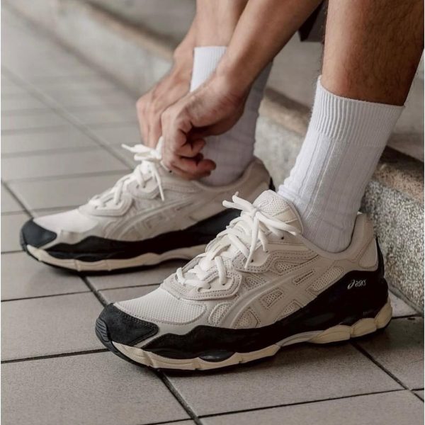 Person tying white ASICS sneakers while sitting on a curb, wearing white crew socks.