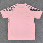 Pink short-sleeve shirt seen from the back, featuring black wing-like patterns on the shoulders laid on a gray carpet.