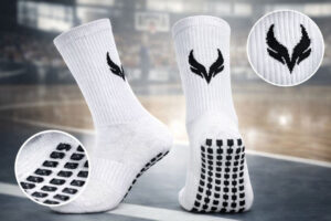 5967537890642300083 | UAE SPORT STORE Pair of white athletic socks with black grip dots on the soles and a logo on the cuff, shown on a gym court surface.