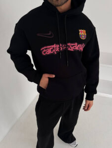 Person wearing a black hoodie with FC Barcelona crest and pink graffiti-style text across the chest, standing in a white room.