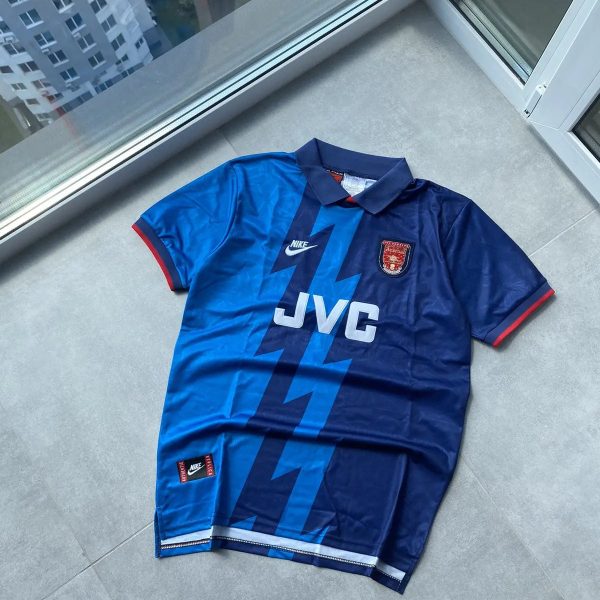 Blue soccer jersey laid flat on a light gray tiled floor beside a glass door, featuring a zigzag lighter blue pattern, Nike logo, JVC sponsor, and team crest on the chest.