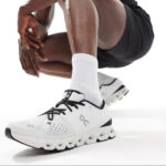 5999176457990638392 | UAE SPORT STORE Close-up of a white running shoe with black laces and a chunky sole on a person wearing white socks.