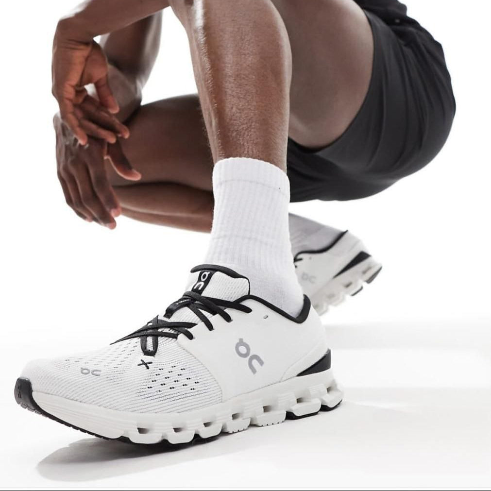 5999176457990638392 | UAE SPORT STORE Close-up of a white running shoe with black laces and a chunky sole on a person wearing white socks.