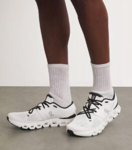 5999176457990638393 | UAE SPORT STORE White athletic running shoes with black laces and mesh detailing, worn with light gray socks on a person standing on a neutral floor.