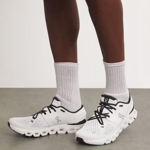 White athletic running shoes with black laces and mesh detailing, worn with light gray socks on a person standing on a neutral floor.