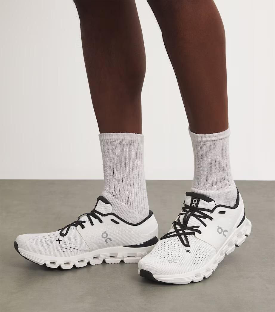 5999176457990638393 | UAE SPORT STORE White athletic running shoes with black laces and mesh detailing, worn with light gray socks on a person standing on a neutral floor.