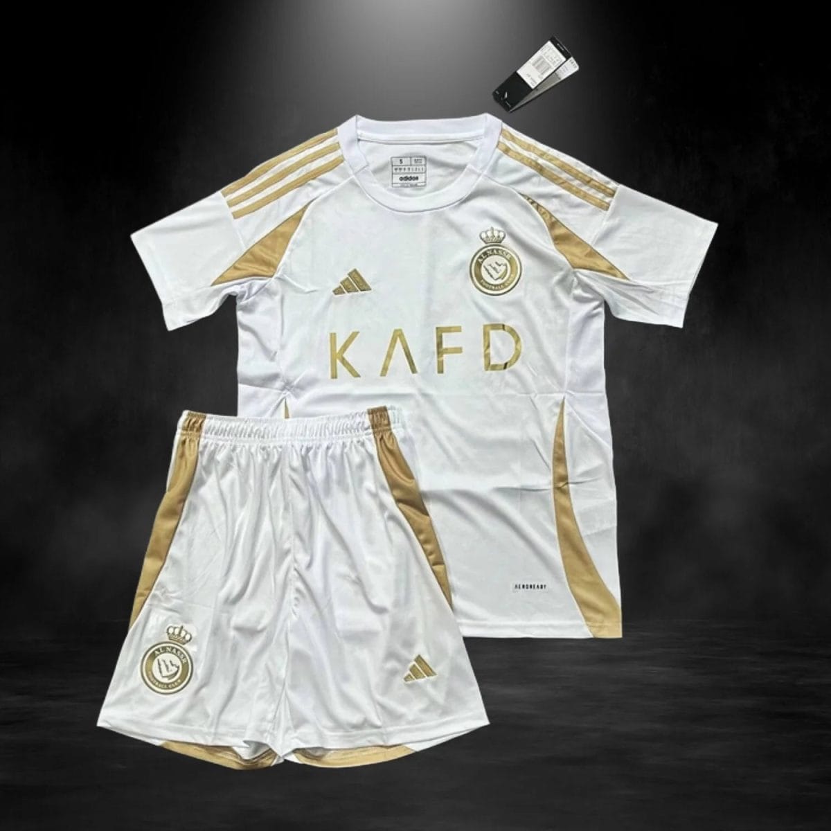 White soccer jersey and shorts with gold trim and crest on a dark background, laid flat for product display.