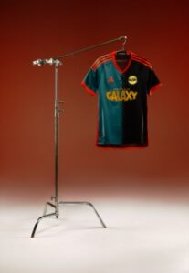 5CQBPVgho4aAkbU | UAE SPORT STORE Soccer jersey on a metal stand in a studio, teal and black with red trim and the word GALAXY on the chest.