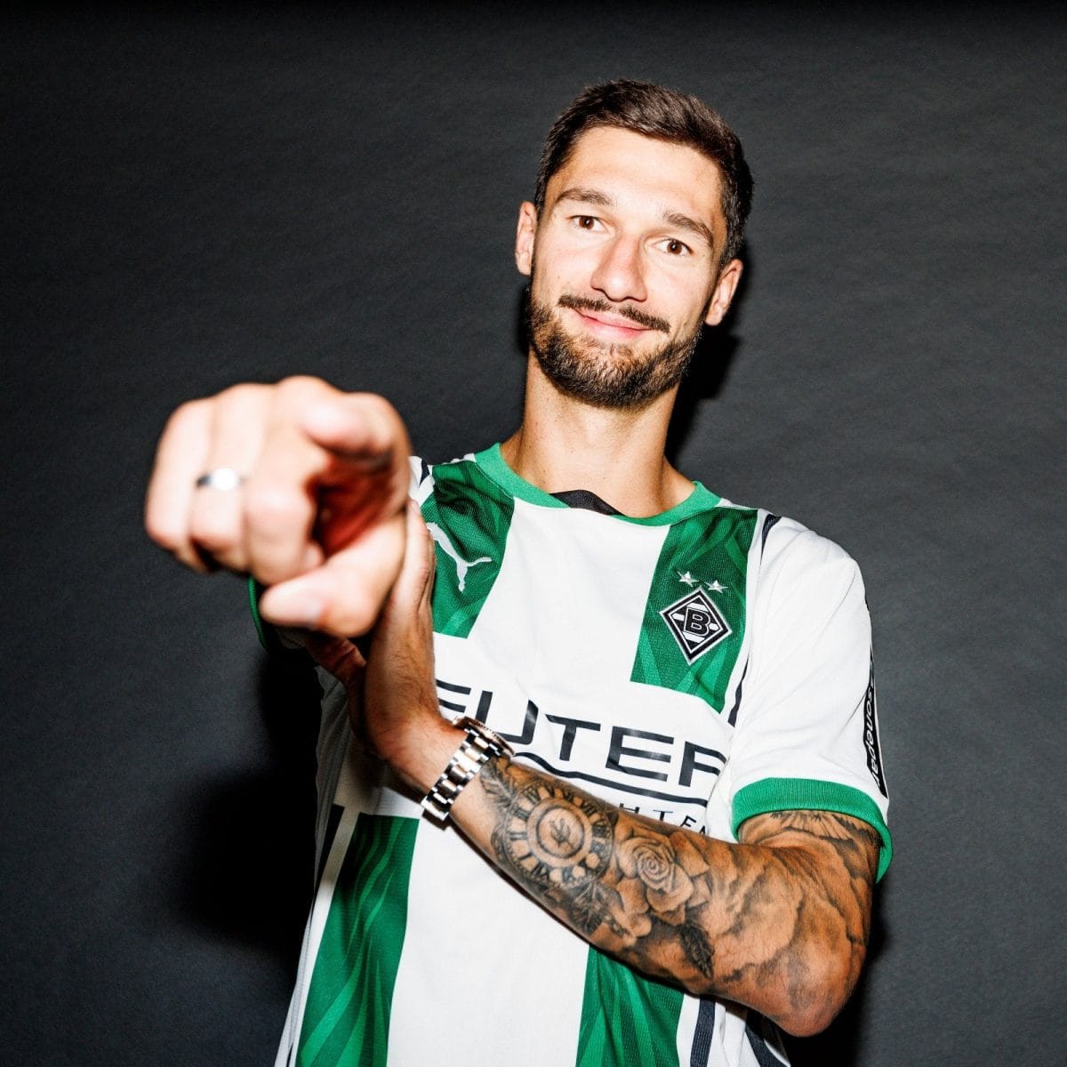 Man in a green-and-white striped soccer jersey points at the camera with a friendly smile.