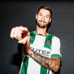 Man in a green-and-white striped soccer jersey points at the camera with a friendly smile.