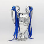 Silver trophy with blue ribbons draped over its handles, standing on a pedestal, symbolizing victory or achievement.