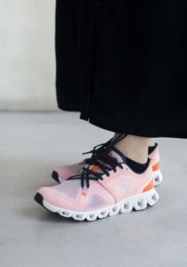 Pink athletic sneakers with black laces and a white segmented sole on a person wearing black pants, standing on a light floor.
