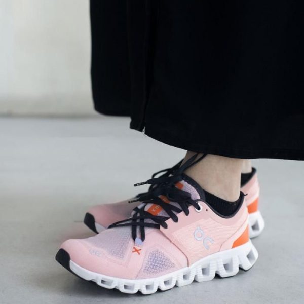 Pink athletic sneakers with black laces and a white segmented sole on a person wearing black pants, standing on a light floor.