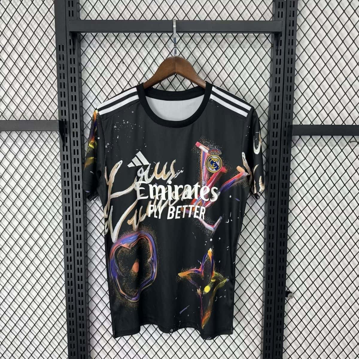 Real Madrid jersey hanging on a wooden hanger against a metal grid, with white stripes on the shoulders and a colorful splash design and Emirates branding.
