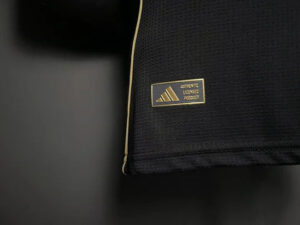 Close-up of a black backpack with beige piping and a gold Adidas patch reading 'AUTHENTIC LICENSED PRODUCT'.
