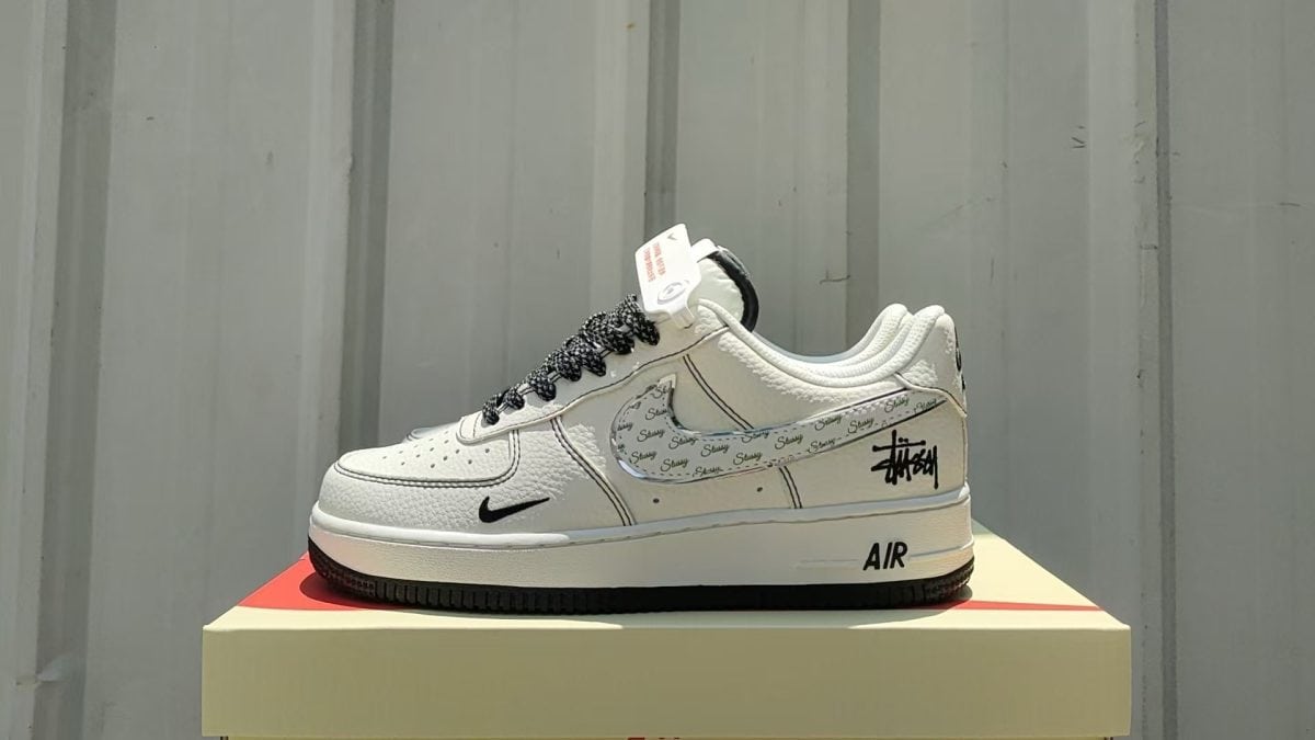 White Nike Air sneakers with a patterned Swoosh and Stussy logo on the side, placed on a beige box against a grey backdrop.